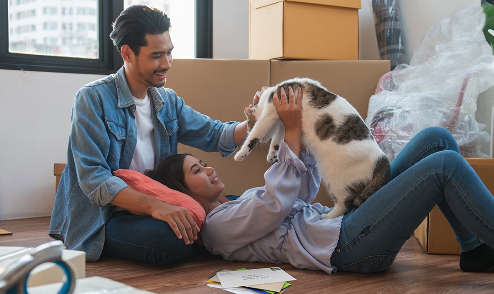 A smiling couple play with a cat on the floor. There is a small stack of mail beside them and stacked cardboard boxes in the background.
