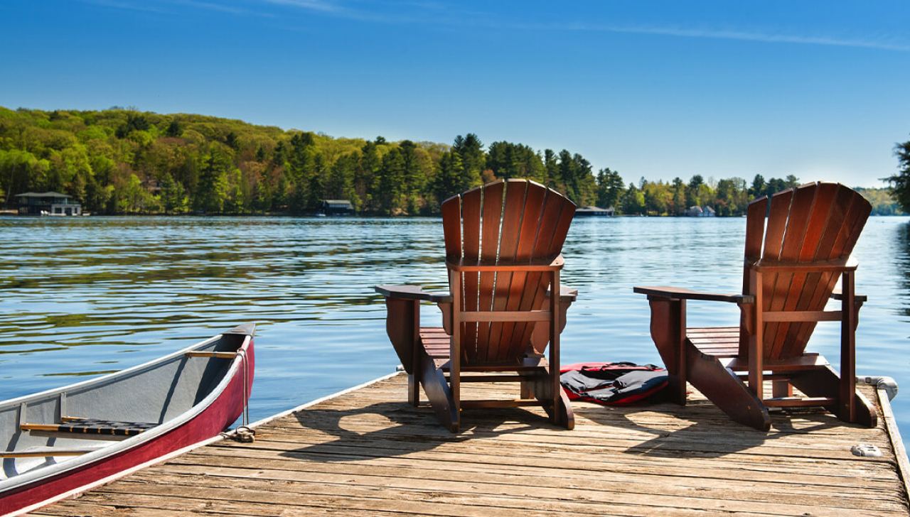 Two wooden chairs sit on a lakeside dock, next to a canoe.