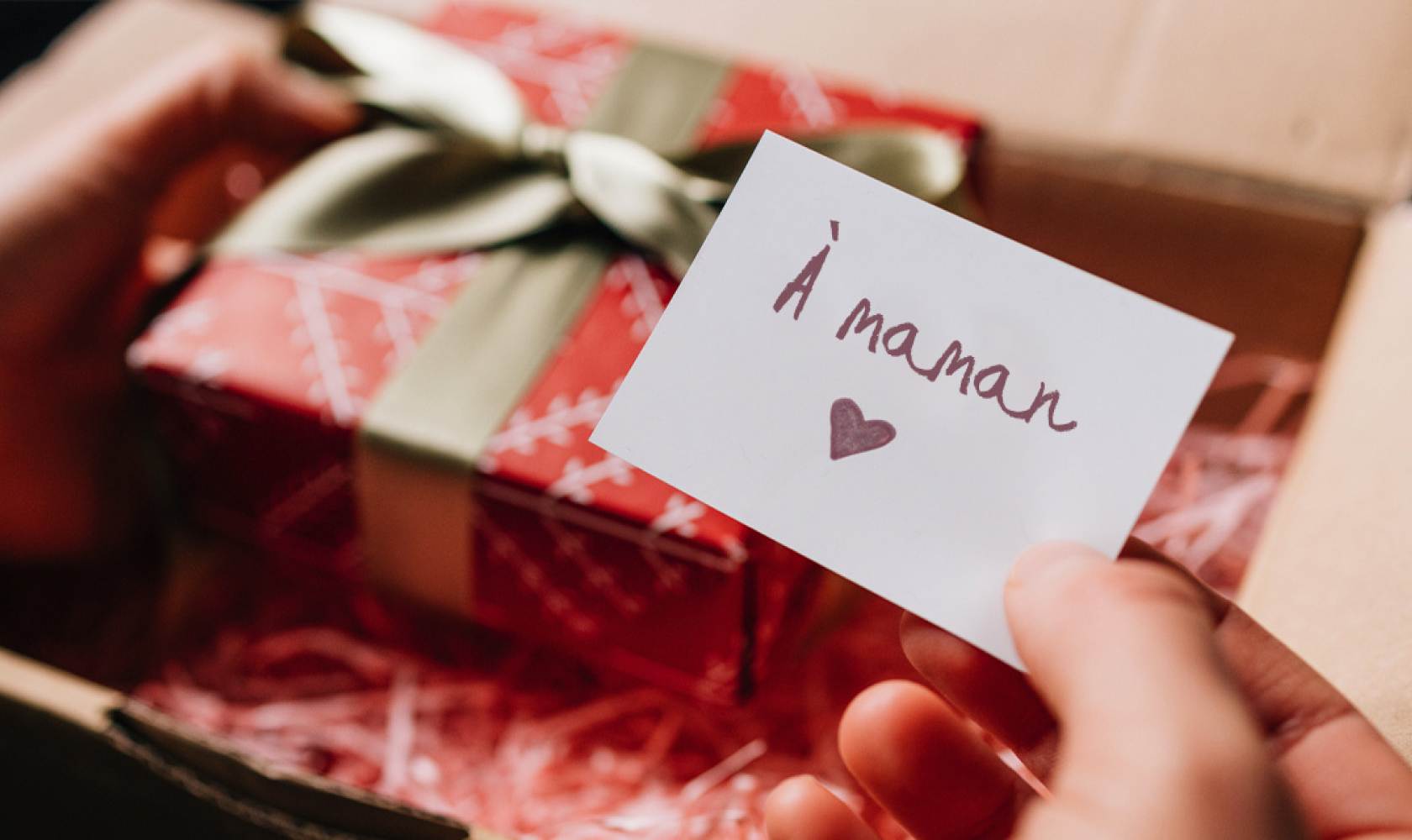 Des mains ouvrent un colis qui contient une boîte-cadeau emballée et une carte sur laquelle il est écrit « À maman »