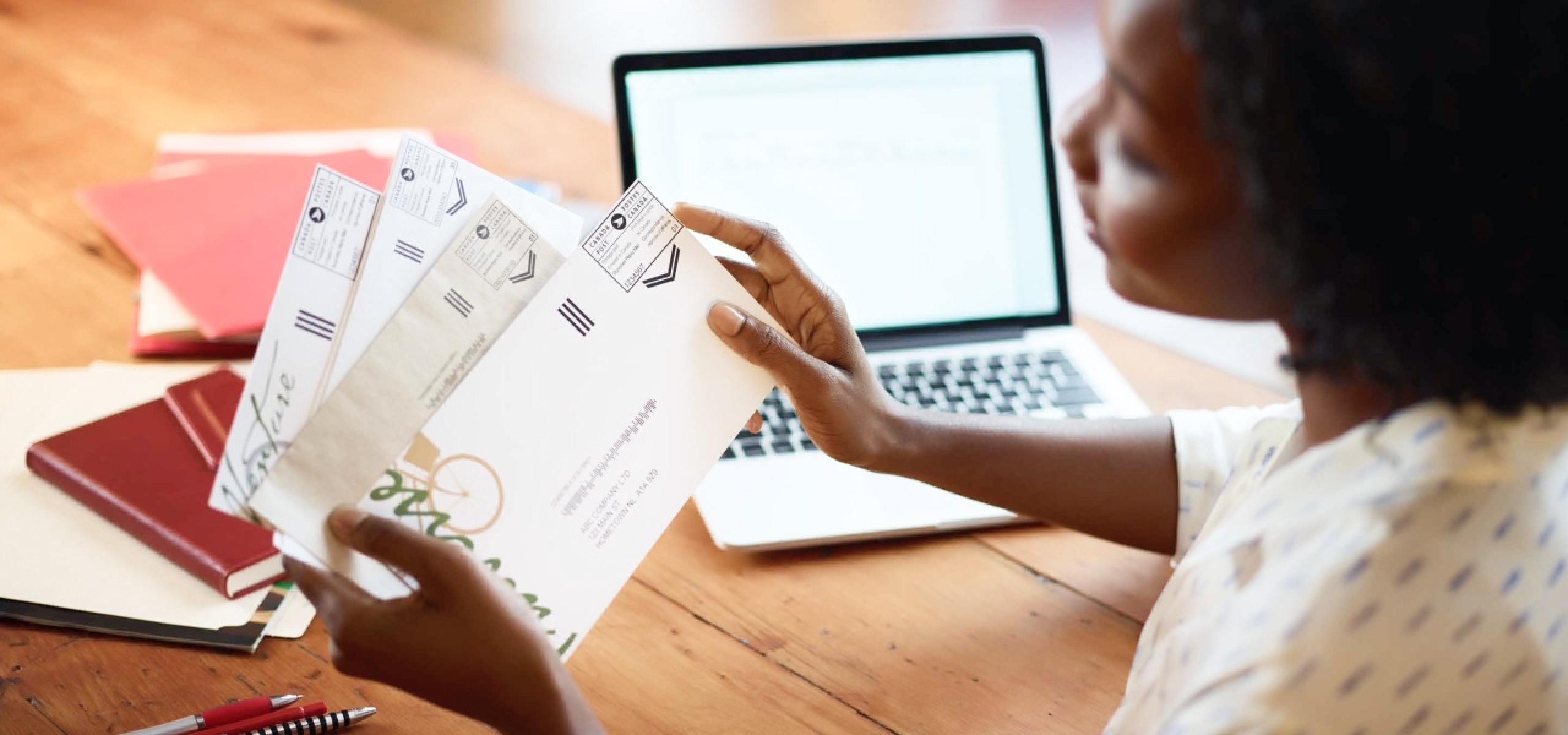 Une femme à son bureau tient du courrier d’affaires. Il y a une vignette postale de Postes Canada dans le coin supérieur droit de chaque enveloppe.